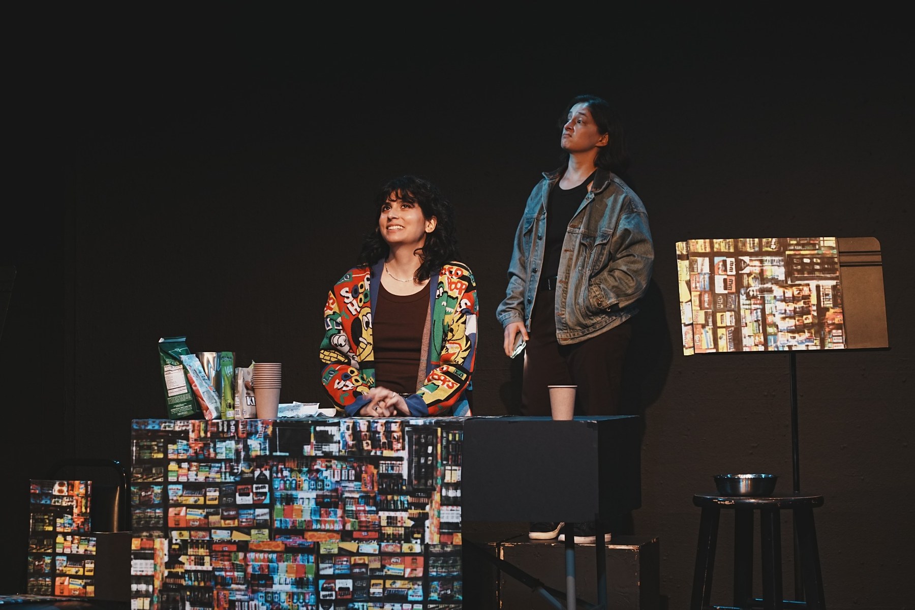 Two performers behind the counter of the bodega set in Bodega Cat; one sits smiling while the other looks upward.