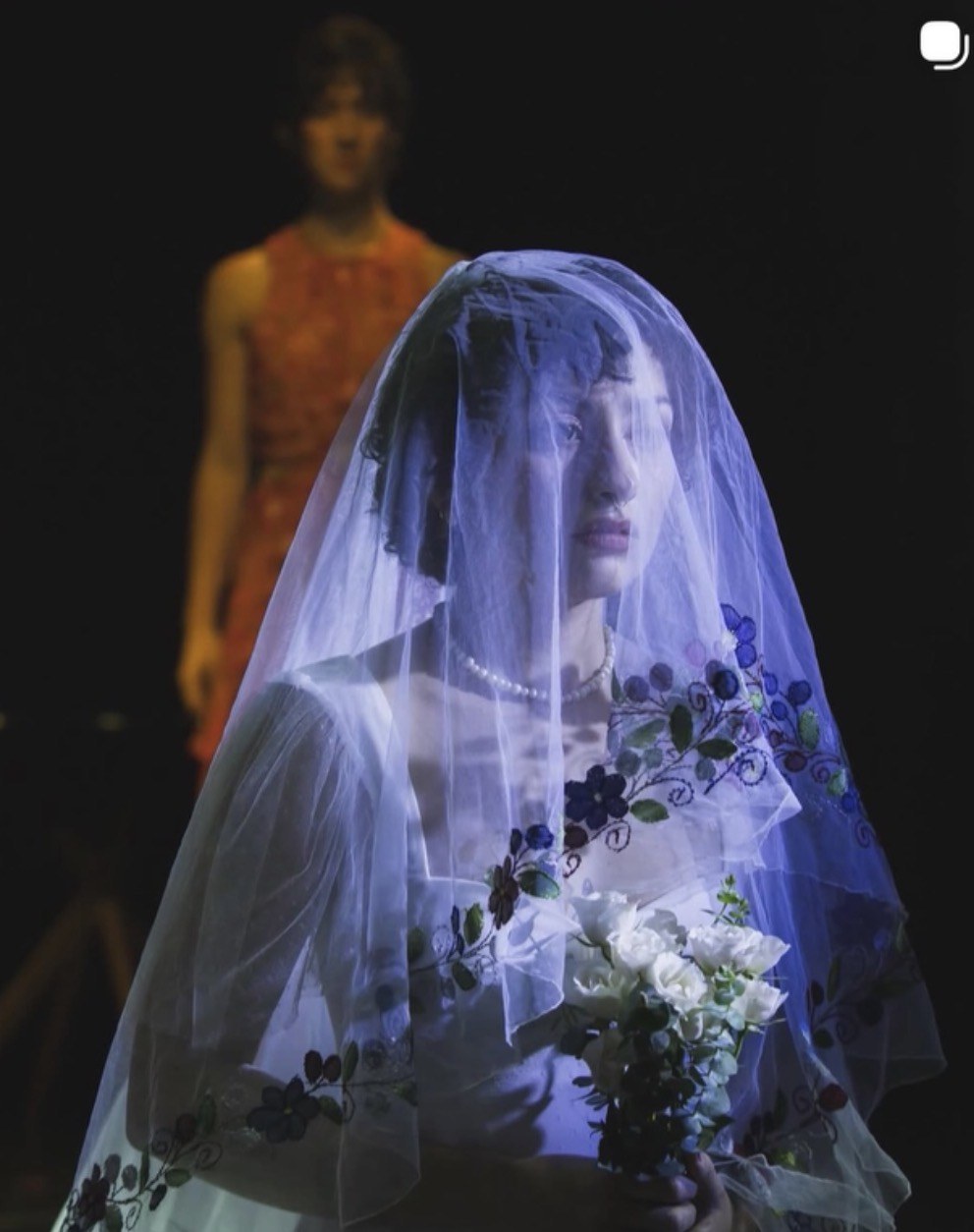 A performer in a bridal veil holds white flowers under blue stage light in Something Blue.